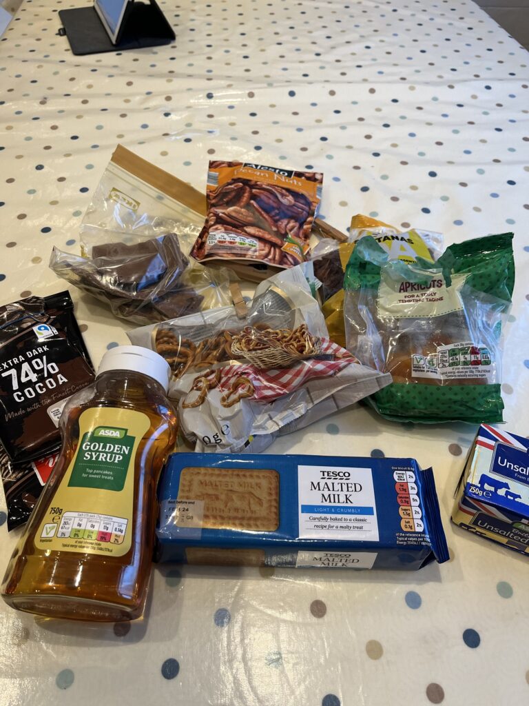 there are ingredients for making rocky road on a table: a bottle of golden syrup, a packet of malted milk biscuits, pretzels, butter, dried fruit and nuts and chocolate